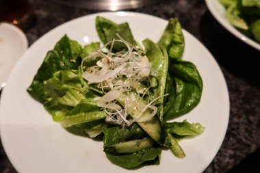 Beyaz bir tabakta servis edilen taze salatanın yanına marul yaprakları, ince dilimlenmiş salatalıklar ve dilimlenmiş soğan serpiştirilmiş..