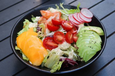 Mango, avokado dilimlenmiş, vişneli domates, marul, lahana ve yengeç etli salatayı açık hava kamp masasında siyah bir masaya koyun.