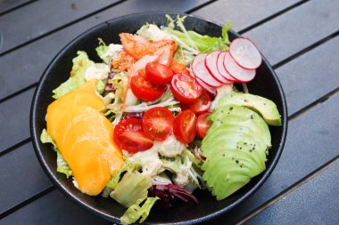 Mango, avokado dilimlenmiş, vişneli domates, marul, lahana ve yengeç etli salatayı açık hava kamp masasında siyah bir masaya koyun.