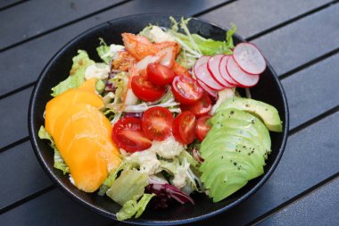Mango, avokado dilimlenmiş, vişneli domates, marul, lahana ve yengeç etli salatayı açık hava kamp masasında siyah bir masaya koyun.