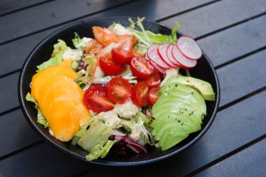 Mango, avokado dilimlenmiş, vişneli domates, marul, lahana ve yengeç etli salatayı açık hava kamp masasında siyah bir masaya koyun.