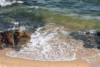 Temiz dalgalar kumlu bir sahile vuruyor, pürüzsüz çakıl taşları ve kayaların kısmen battığını gösteriyor..