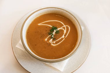 Süslü kremalı balkabağı çorbası ve beyaz masa örtüsü kaplı bir tabağa yerleştirilmiş beyaz bir kase içindeki bir bitki serpintisi..