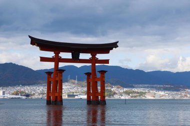 Miyajima, Hiroşima, Japonya - 9 Aralık 2024: Bulutlu bir günde kayalar ve deniz yosunlarını gösteren sığ suyla gündüz vakti Itsukushima Tapınağı 'nın Torii Kapısı' nda yüzen kırmızı shinto