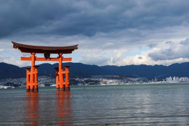Miyajima, Hiroşima, Japonya - 9 Aralık 2024: Bulutlu bir günde kayalar ve deniz yosunlarını gösteren sığ suyla gündüz vakti Itsukushima Tapınağı 'nın Torii Kapısı' nda yüzen kırmızı shinto