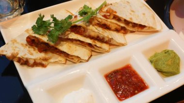 Hint quesadillasını yakın plan üçgenlere ayırın, güzelce yerleştirin ve taze kişniş ile süsleyin, üç soslu sos, kırmızı salsa, ekşi krema ve yeşil guacamole ile servis edin.