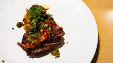 Çok güzel bir tabaktaki bifteğin dış tarafı koyu kavrulmuş, üstü doğranmış domates, otlar ve kapari ile kaplı beyaz tabakta fotokopi alanı ile kapari ile kaplanmış.