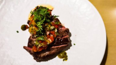 Çok güzel bir tabaktaki bifteğin dış tarafı koyu kavrulmuş, üstü doğranmış domates, otlar ve kapari ile kaplı beyaz tabakta fotokopi alanı ile kapari ile kaplanmış.