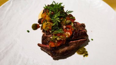 Çok güzel bir tabaktaki bifteğin dış tarafı koyu kavrulmuş, üstü doğranmış domates, otlar ve kapari ile kaplı beyaz tabakta fotokopi alanı ile kapari ile kaplanmış.