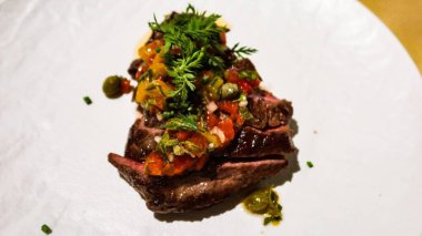 Çok güzel bir tabaktaki bifteğin dış tarafı koyu kavrulmuş, üstü doğranmış domates, otlar ve kapari ile kaplı beyaz tabakta fotokopi alanı ile kapari ile kaplanmış.