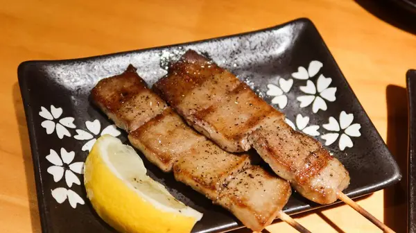 Izakaya 'da limon dilimli fotokopi alanı olan siyah bir tabakta iki tane ızgara öküz dili şiş.