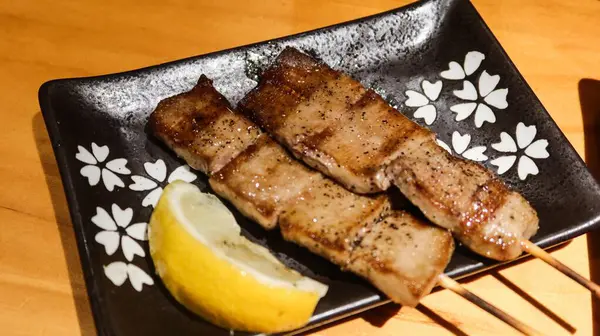 Izakaya 'da limon dilimli fotokopi alanı olan siyah bir tabakta iki tane ızgara öküz dili şiş.