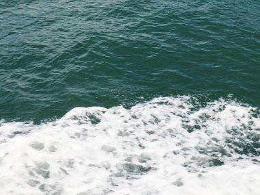 Yüzeyde beyaz köpüklü derin yeşil okyanus dalgalarının dinamik bir görüntüsü, bir teknenin perspektifinden çekilmiş, denizin gücünü ve hareketini gösteriyor..