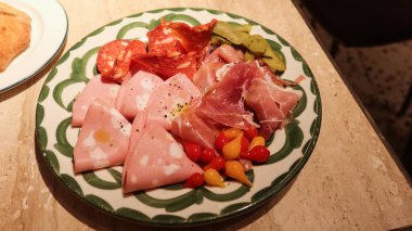 Salam, prosciutto ve mortadella da dahil olmak üzere, ahşap bir masada yeşil çerçeveli dekoratif bir tabakta servis edilen turşu turşusu ve kiraz biberiyle süslenmiş jambon tabağının hemen yanında.
