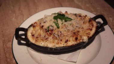 Erimiş peynir ve taze maydanozla kaplı, siyah dökme demir bir tavada servis edilen peynirli makarna ve peyniri, sıcak mermer masa arkaplanlı basit beyaz bir tabakta sunuluyor.