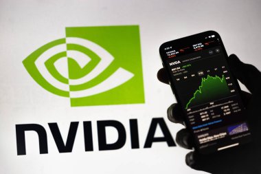 Ters ışıklandırılmış bir el, Apple borsa uygulamasında yükselen yeşil Nvidia hisse senedi fiyatlarını gösteren bir telefon tutuyor. Arkaplanda logonun bulanık bir ekranı var.