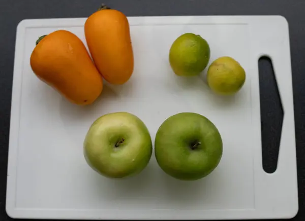 İki hindistan cevizi, iki misket limonu ve iki elma, siyah arka planda beyaz bir kesim tahtasında.