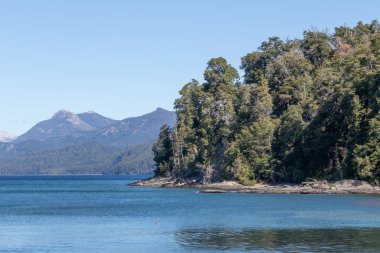 Güneşli bir günde, Arjantin, Lago Nahuel Huapi 'de, açık mavi gökyüzü ve gölün sularının sakin dalgalarıyla çevrili, bitki ve kayalarla dolu dağların yatay fotoğrafı.