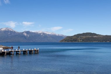 Kenarında bir rıhtım bulunan devasa sakin bir gölün yatay fotoğrafı ve hafif sis ile kaplı dağlarla dolu bir yer ve Arjantin, Lago Nahuel Huapi 'de az miktarda bulutlu açık mavi bir gökyüzü.