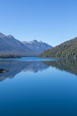 Gökyüzünü yansıtan bir göl manzarasının rahatlatıcı duvar kağıdı, bitki örtüsü dolu dağ sıraları, ve bitkiler ve ağaçlarla çevrili.