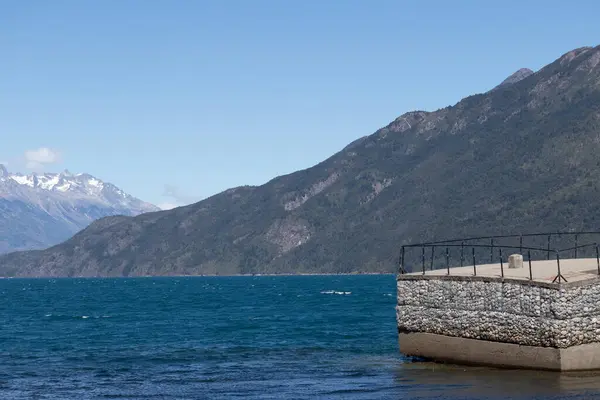 Lago Pueblo 'da kayalık bir sahilden bir gölün suyuna kayalar, bitkiler ve muazzam büyüklükte bir dağ sırasının yatay fotoğrafı.