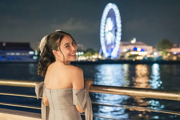 Happy beautiful young asian woman in dress smiling and sightseeing illuminated ferris wheel during cruise ship on the river in the night at Bangkok