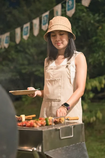 Young asian woman grilling barbeque skewers meat on bbq grill in backyard on picnic time at night
