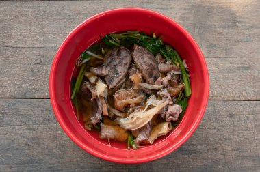Combination beef and offal noodle soup with vegetable in red bowl is Thai Asian food on wooden table in local restaurant