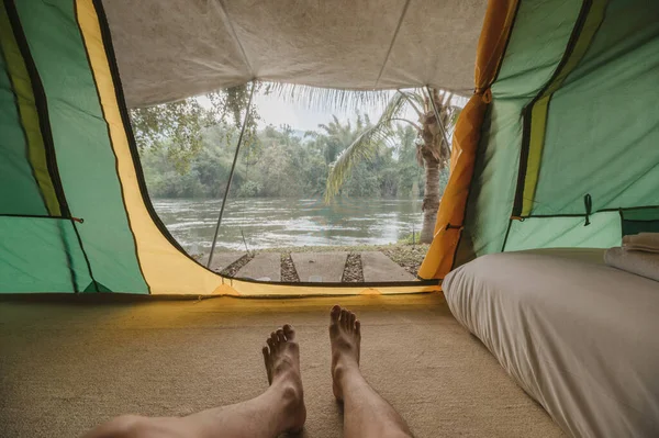 Legs of man relaxing inside a comfortable camping tent on campground in the tropical forest by riverside on summer vacation