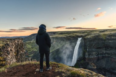 Rear view of man in winter coat standing and looking at Majestic Haifoss waterfall flowing in volcanic canyon among the Icelandic Highlands on summer at Iceland