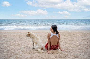 Güzel, genç Asyalı bir kadın, tatilde tropikal denizde bir köpekle sahilde oturuyor. Yaz ve tatil kavramı