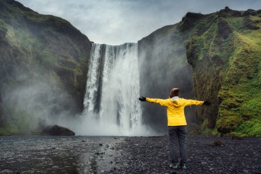 Sarı ceketli genç Asyalı kadın turist yaz mevsiminde İzlanda 'da uçurumdan akan Skogafoss şelalesinin tadını çıkarıyor. İzlanda 'nın ünlü yeri, seyahat yeri