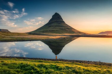 Görkemli simetri volkanı Kirkjufell 'in göl yansıması ve erkek turistin yaz sabahı İzlanda' daki Snaefellsnes yarımadasında durduğu hava manzarası.