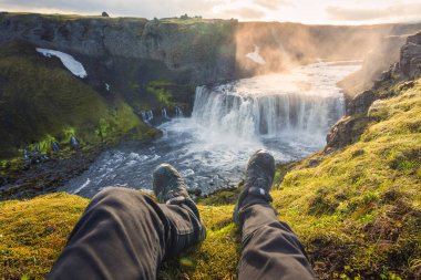 Akşamları İzlanda 'nın dağlarında hızla akan Axlafoss şelalesi ile volkanik dağın kenarında oturan botlar giyen adamın bacakları kişisel perspektifi.