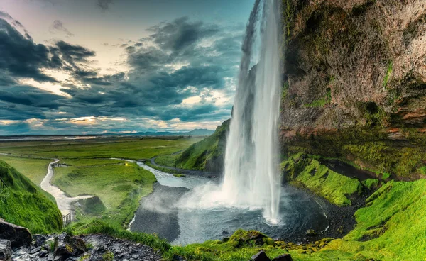 Güzel Seljalandsfoss şelalesinin panoramik manzarası yazın İzlanda 'da kasvetli bir günde ve yemyeşil bir tarlada akıyor.