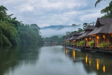 Kanchanaburi, Tayland 'da kasvetli bir günde Kwai Nehri' nde yüzen güzel ahşap bir tatil köyü ve sisli bir dağ.