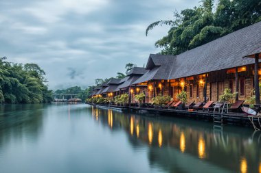 Kanchanaburi, Tayland 'da kasvetli bir günde Kwai Nehri' nde yüzen güzel ahşap bir tatil köyü ve sisli bir dağ.