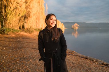 Mutlu güzel Asyalı kadın güneş ışığıyla eğleniyor ve gülümsüyor Wanaka Gölü, Yeni Zelanda 'daki sarı sonbahar ağaçlarında parlıyor.