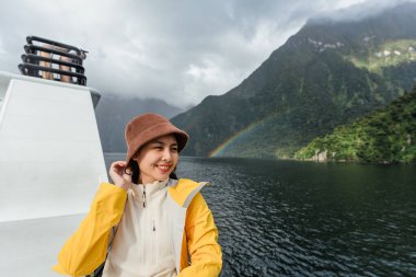 Sarı ceketli Asyalı kadın Milford Sound, Yeni Zelanda 'daki göl kıyısındaki fiyortta dağ ve gökkuşağıyla eğleniyor.