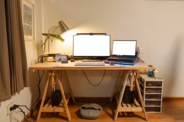 Bilgisayarı, monitörü, bilgisayar aygıtı, aydınlatma lambası, ahşap masa üzerindeki yeşil yaprak ve evdeki ergonomik sandalyeli fotoğrafçının iç işyeri.