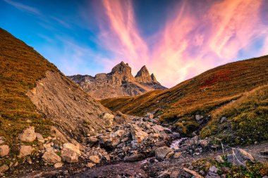 Dramatic landscape of sunset sky over Aiguilles d Arves with iconic three rocky mountain peak and cascade flowing in French Alps valley at Savoie, France clipart