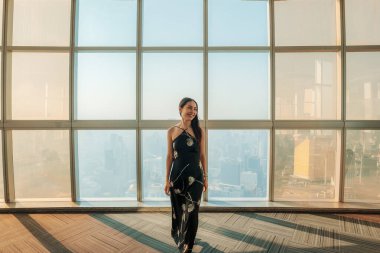 Siyah elbiseli güzel Asyalı kadın gözlem güvertesinden manzaranın tadını çıkarıyor ve şehir merkezindeki Bangkok gökdeleninin modern cam penceresinden.