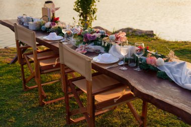 Gün batımında ahşap uzun masa, sandalye, sofra takımı, çatal bıçak takımı, mum ve flora dekorasyonuyla kırsal ve romantik bir açık hava yemek düzeni. 