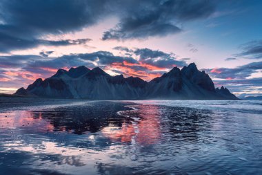Yazın Stokksnes yarımadasında, İzlanda 'da volkanik Vestrahorn Dağı' nın tepesindeki dramatik günbatımı manzarası kara kumları yansıtıyor.