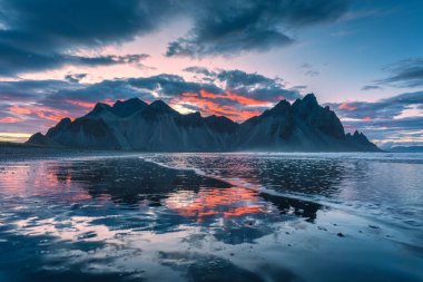 Yazın Stokksnes yarımadasında, İzlanda 'da volkanik Vestrahorn Dağı' nın tepesindeki dramatik günbatımı manzarası kara kumları yansıtıyor.