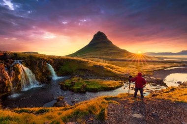 Kırmızı ceketli bir fotoğrafçı Kirkjufellsfoss ile birlikte Kirkjufell Dağı 'nın üzerinde parlak bir gündoğumuyla karşı karşıya. Sonbaharda Snaefellsnes yarımadasında.
