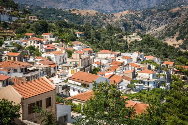 A vertical closeup shot of buildings with a mountain covered with green trees in the background on a sunny day, Argiroupoli village in Crete, Greece