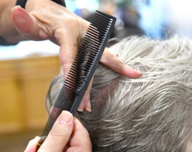 Güzellik uzmanı, yaşlı bir müşterinin saçını kesmeye hazırlanırken elinde tarak tutuyor. Tarağı siyah..