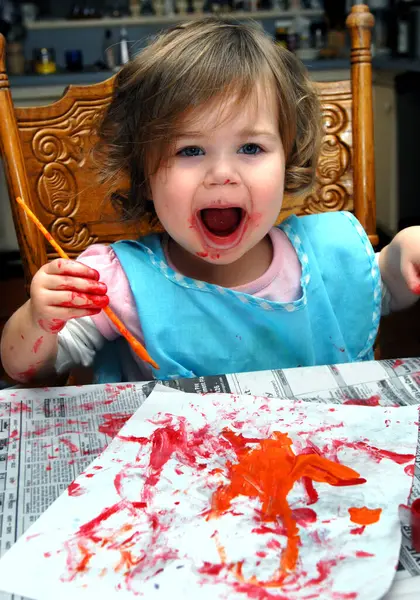 stock image Toddler squaels with joy as she admires her creation.  She is painting with a brush onto a white sheet of paper.  Red paint is smeared around her mouth and on her hands.