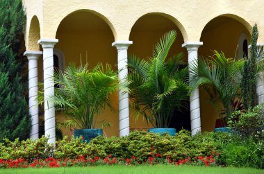 Güzel manzaralı kemerli verdanzada beyaz sütunlar ve sarı boyalı sıva var. Büyük saksı bitkileri verandanın kenarında otururlar. Önplanda kırmızı çiçekler açar.